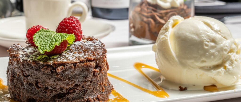 A chocolate lava cake topped with raspberries and mint sits beside a scoop of vanilla ice cream on a white plate, with caramel drizzle and a blurred dessert in the background.