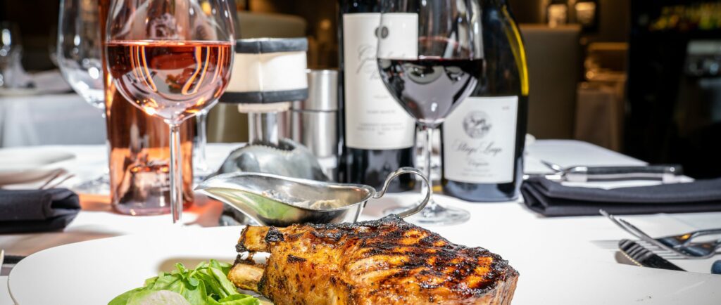 Grilled pork chop on a white plate with greens, a sauce boat, and glasses of red and rosé wine on a set dining table.