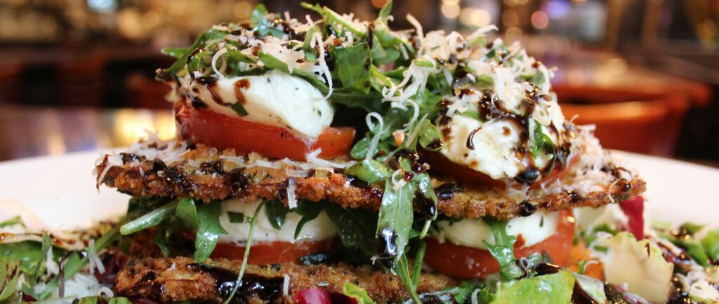 A close-up of a layered salad with fried eggplant slices, tomato, mozzarella, arugula, grated cheese, and a drizzle of balsamic glaze.