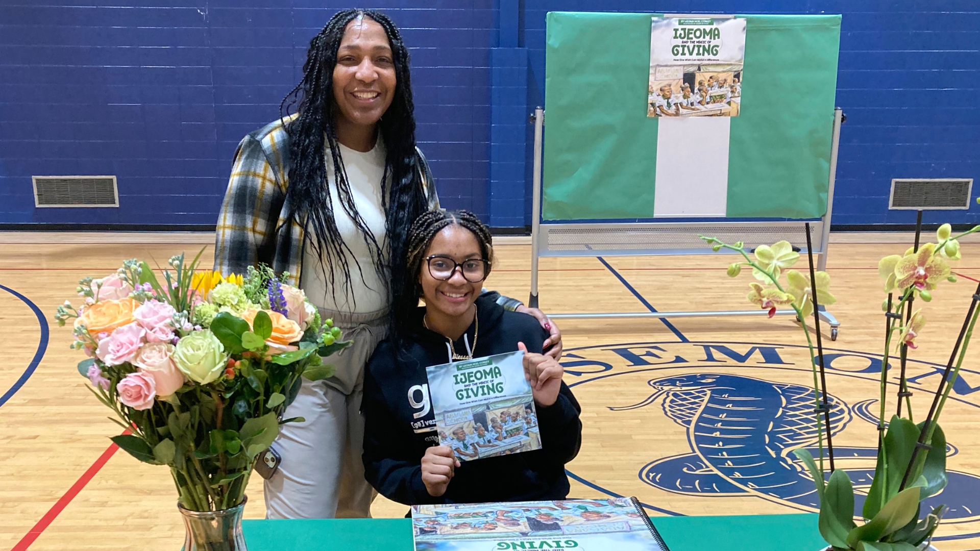Two women pose at a table with flowers and a book titled “Ideoma Giving” in a gymnasium. One sits holding the book while the other stands beside her, both smiling at the camera.