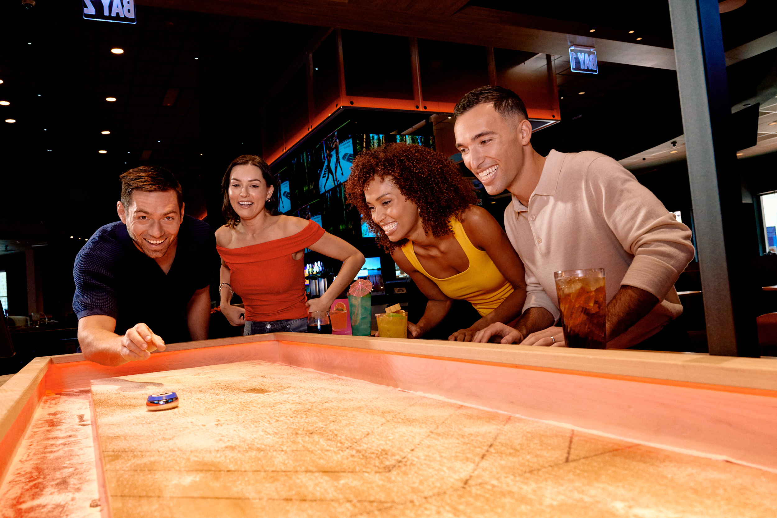 Four people stand around a shuffleboard table in a bar, smiling and watching as one person slides a puck, with drinks on the table.