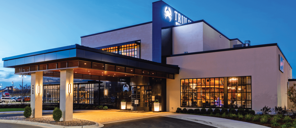 Modern restaurant exterior at dusk with large windows, illuminated signage, a covered entrance, and landscaped surroundings.