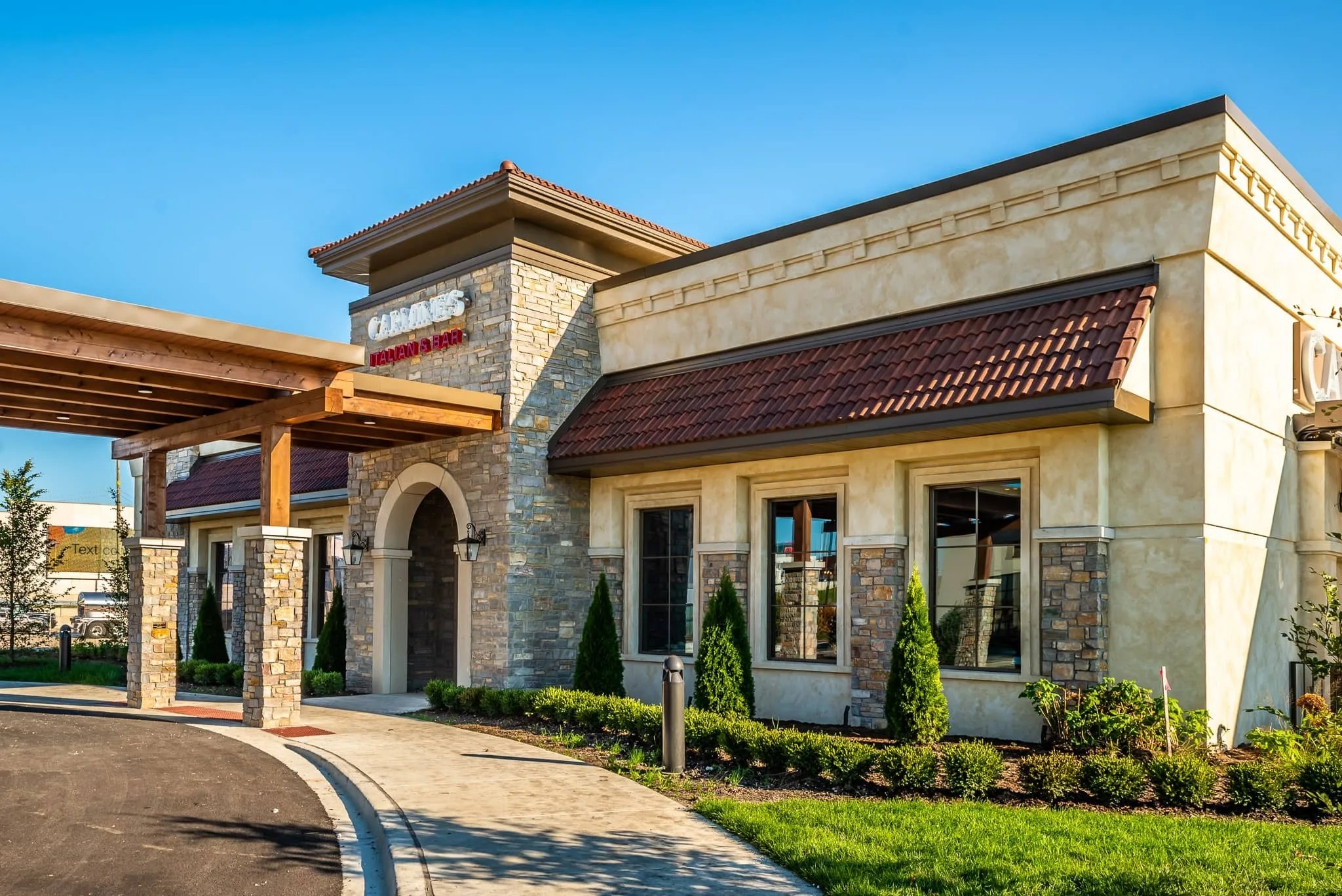 A single-story restaurant building with tan walls, stone accents, red-tiled roof, and manicured landscaping under a clear blue sky.