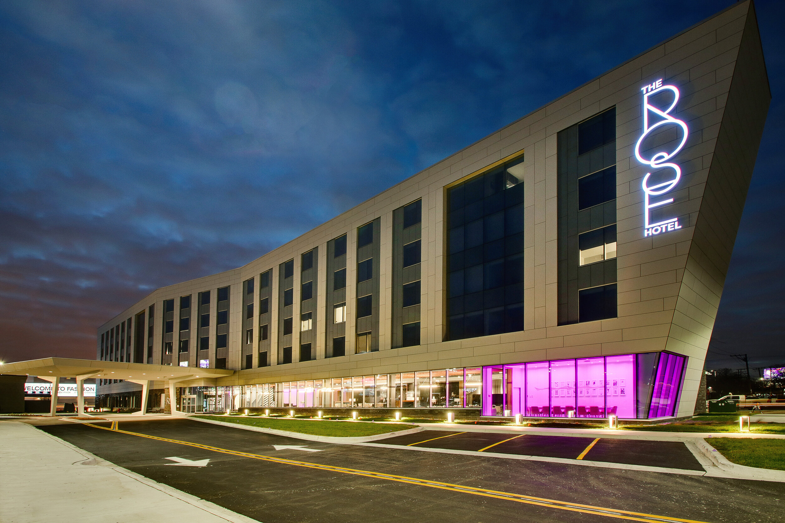 Modern hotel building with angular design, lit by purple and white lights at dusk, with The Rose Hotel sign visible on the exterior.