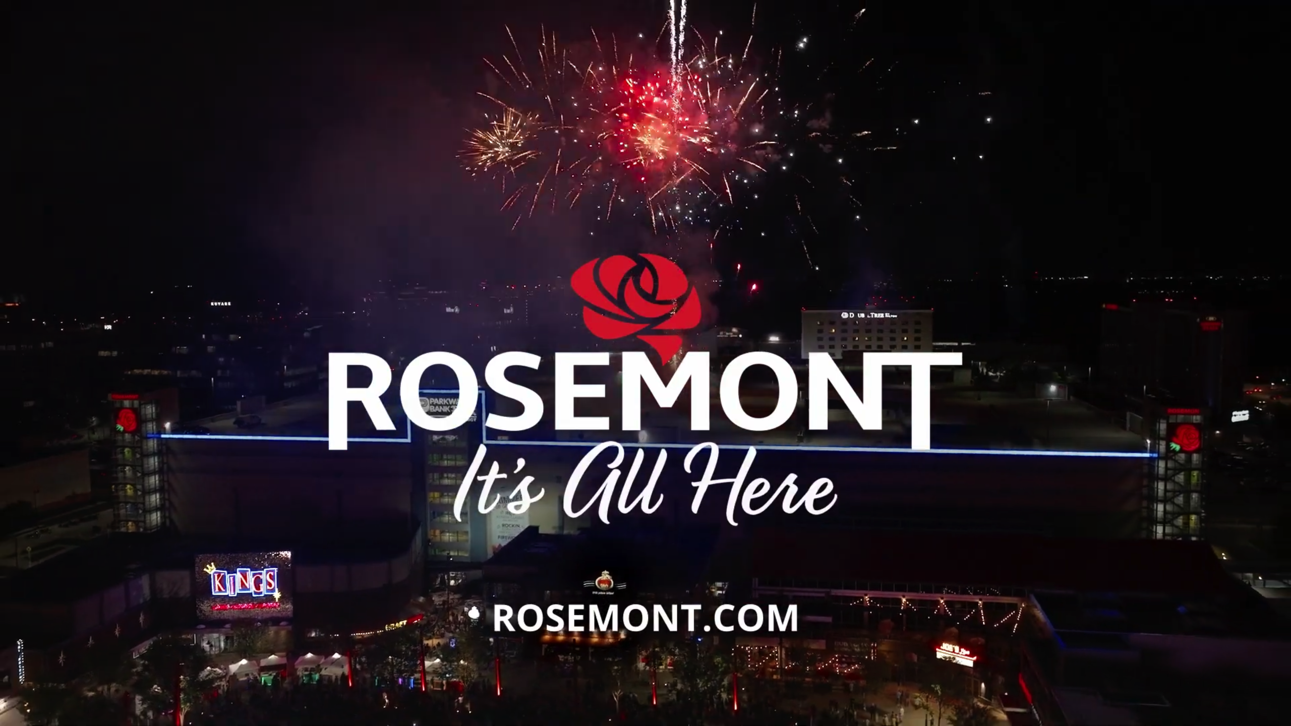 Aerial night view of Rosemont with fireworks in the sky; text reads ROSEMONT Its All Here and ROSEMONT.COM over a cityscape with illuminated buildings.