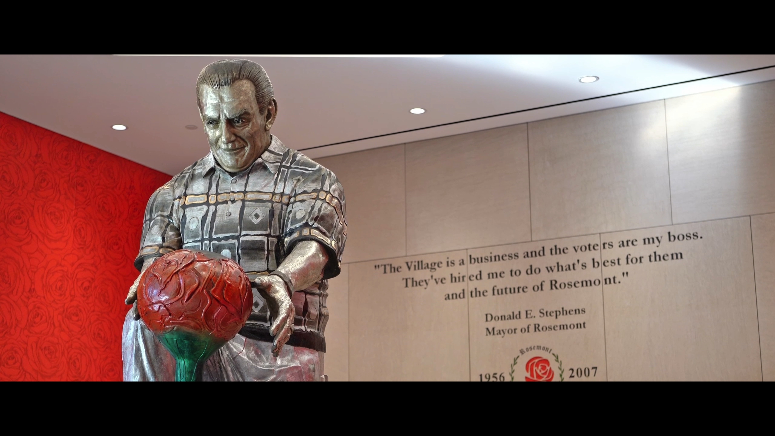 A statue of a man in a patterned shirt holding a red rose stands in front of a wall with a quote from Donald E. Stephens, Mayor of Rosemont, and a memorial plaque.