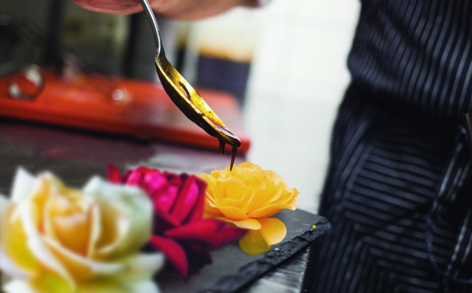A person drizzles sauce from a spoon onto a yellow edible flower on a plate, with other colorful flowers nearby.