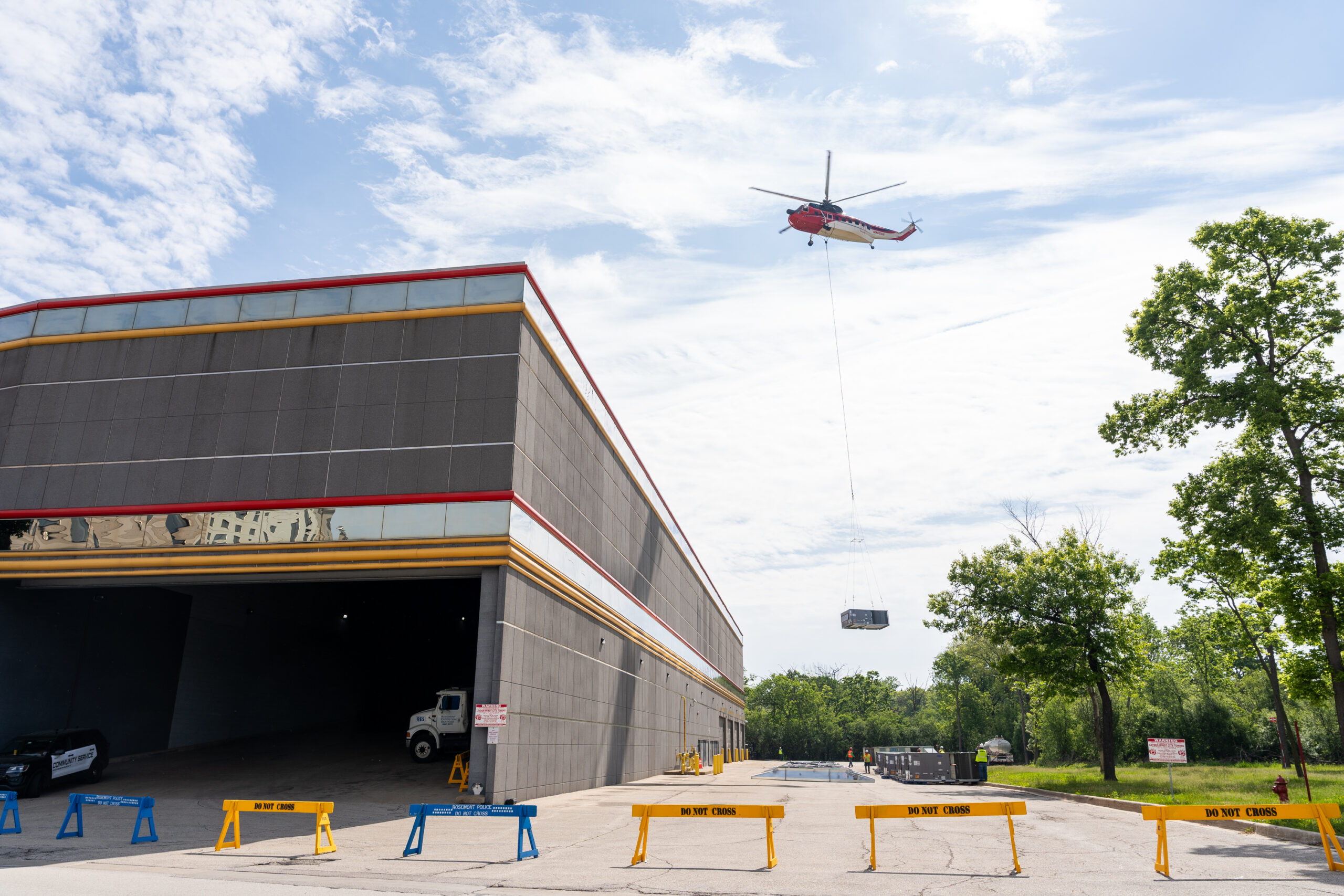 A red helicopter transports a load suspended by a cable above a commercial building, while barricades block off the area below.