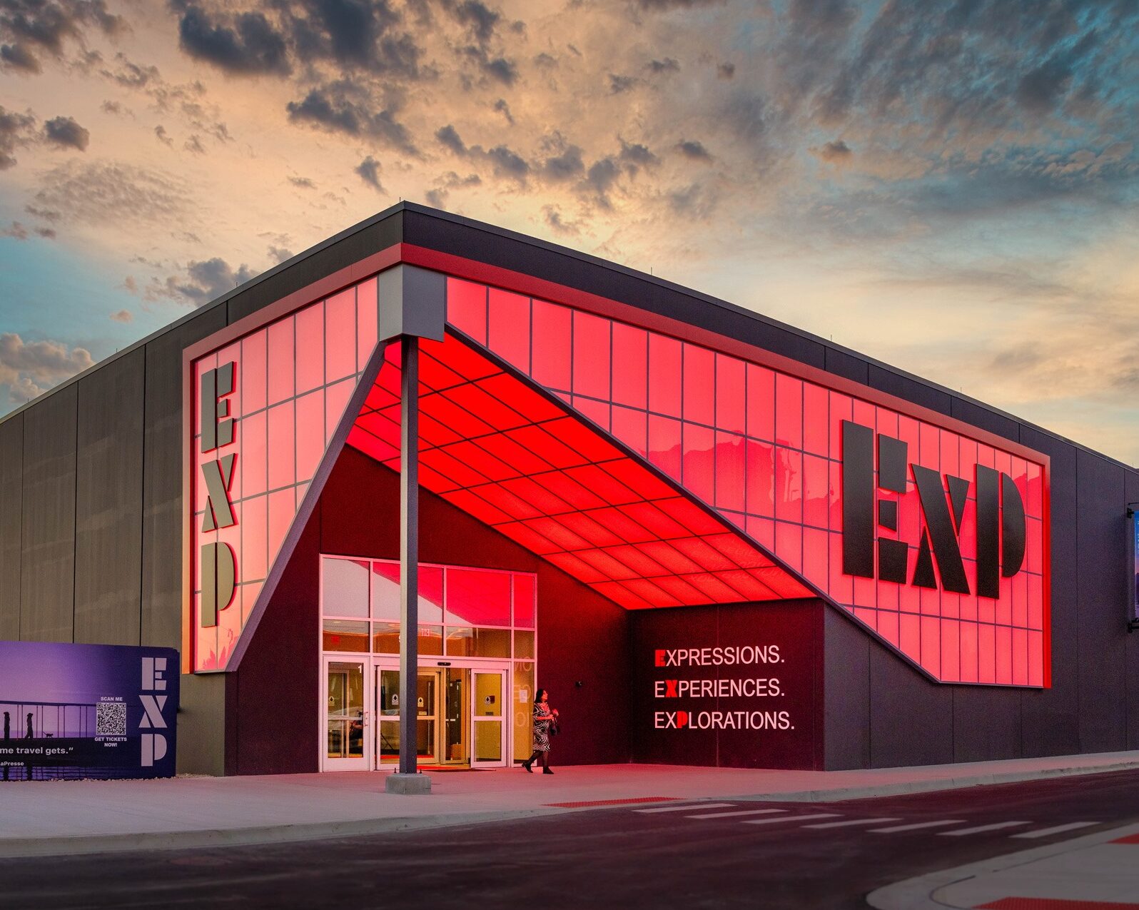 Modern building with a red-lit glass facade displaying the letters “EXP” and text reading “Expressions. Experiences. Explorations.” Two people walk near the entrance at dusk.