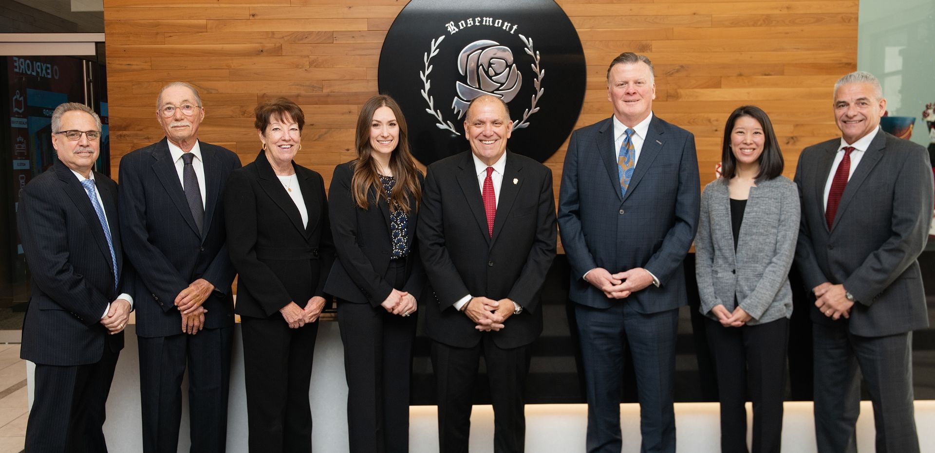 Eight professionally dressed individuals stand in a row in front of a wooden wall and a black and white emblem featuring a rose and laurel wreath.