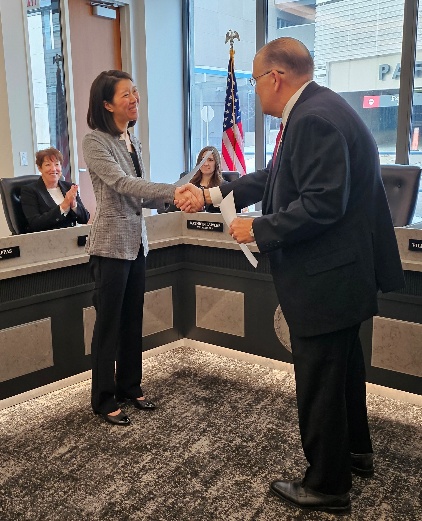 A woman and a man shake hands in a meeting room while two seated women observe; an American flag is visible in the background.