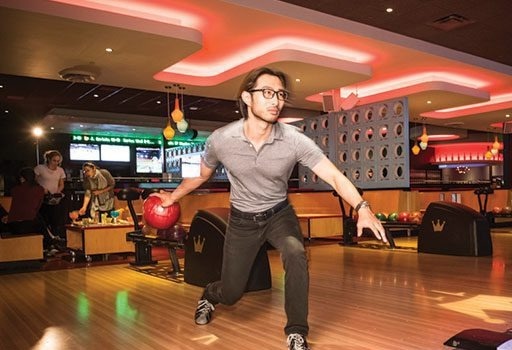 A person in glasses and a gray shirt is bowling, mid-release of a red bowling ball in a brightly lit bowling alley.