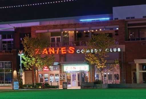 Exterior of Zanies Comedy Club at night with illuminated neon signs and trees in front of the building.