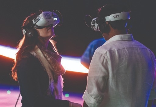 Two people wearing virtual reality headsets and headphones stand facing each other indoors, with blurred lights in the background.