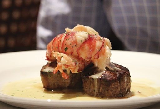 A lobster tail served on top of two filet mignon steaks, covered in a creamy sauce, presented on a white plate.