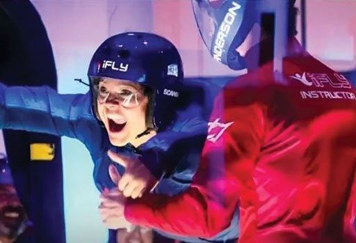 A person in flight gear and helmet is indoor skydiving with an instructor, giving a thumbs up inside a wind tunnel at iFLY.