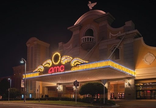 Night view of an illuminated AMC movie theater entrance with marquee lights and architectural details, including a small statue on top of the building.