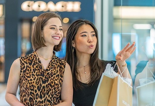 Two women stand in a shopping mall, smiling and looking at a store display, while holding shopping bags.