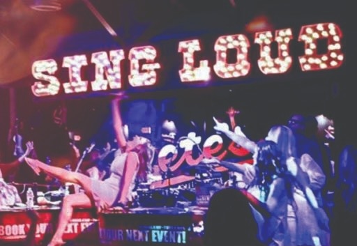 People on stage singing and dancing under a large illuminated SING LOUD sign at a lively indoor music event.
