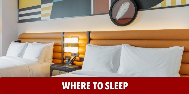 Two neatly made beds with white linens in a modern hotel room, nightstand with lamp between them, and abstract artwork above. Text: WHERE TO SLEEP.