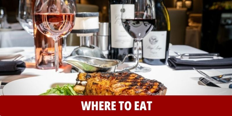 A grilled steak served on a white plate with wine glasses and bottles in the background, on a formal dining table; text reads “WHERE TO EAT.”.