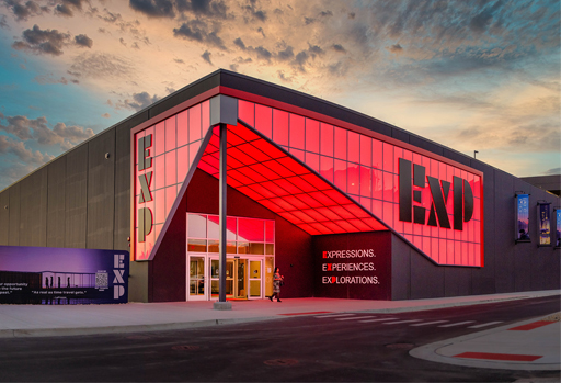 Modern building with a red-lit geometric facade displays the letters EXP and the words “Expressions. Experiences. Explorations.” at dusk. A person stands near the entrance.