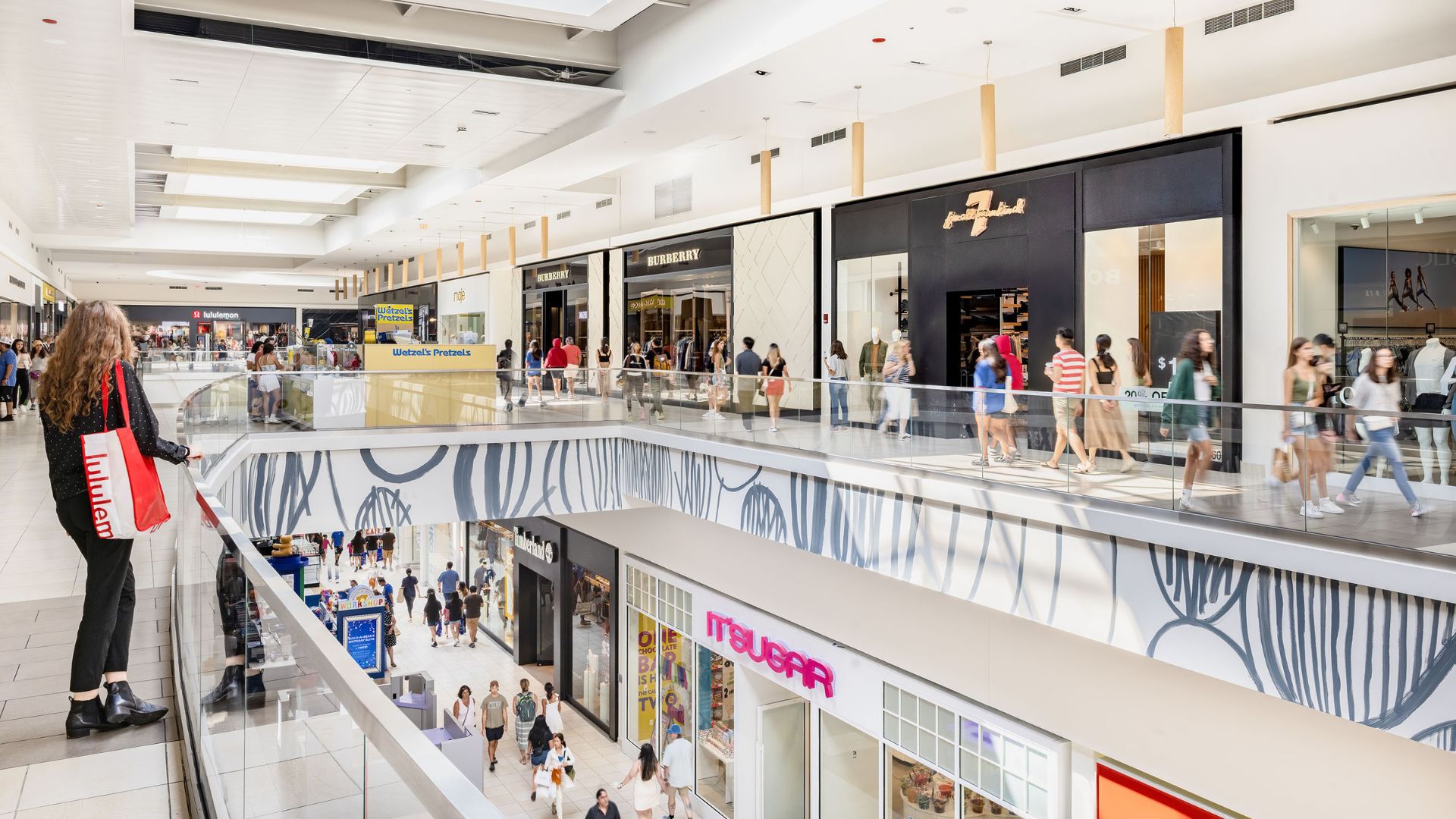 Shoppers walk and browse stores on both levels of a modern indoor shopping mall with glass railings and bright lighting.