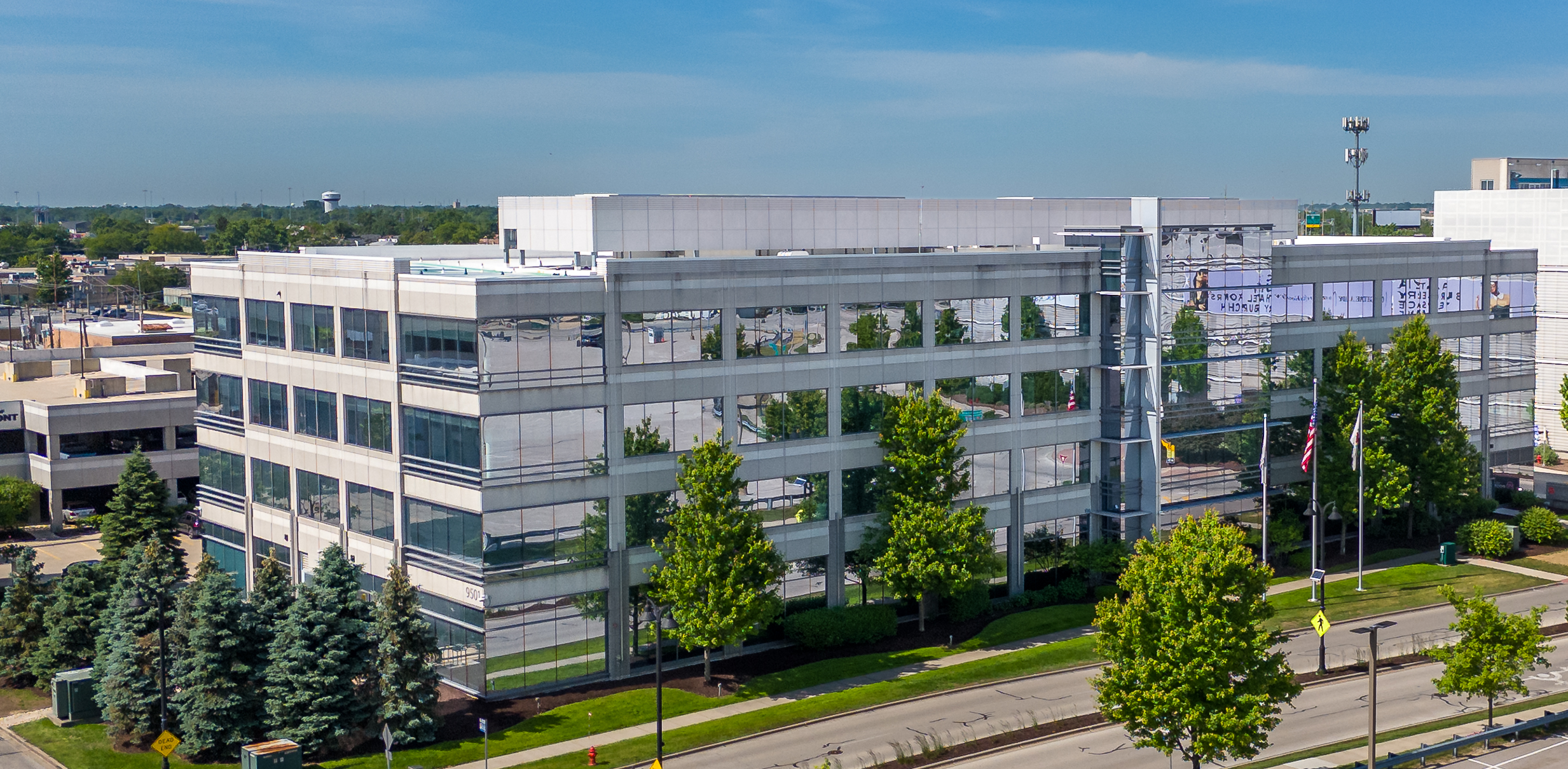 Four-story modern office building with glass exterior, surrounded by trees and landscaping, adjacent to a road with visible traffic markings.