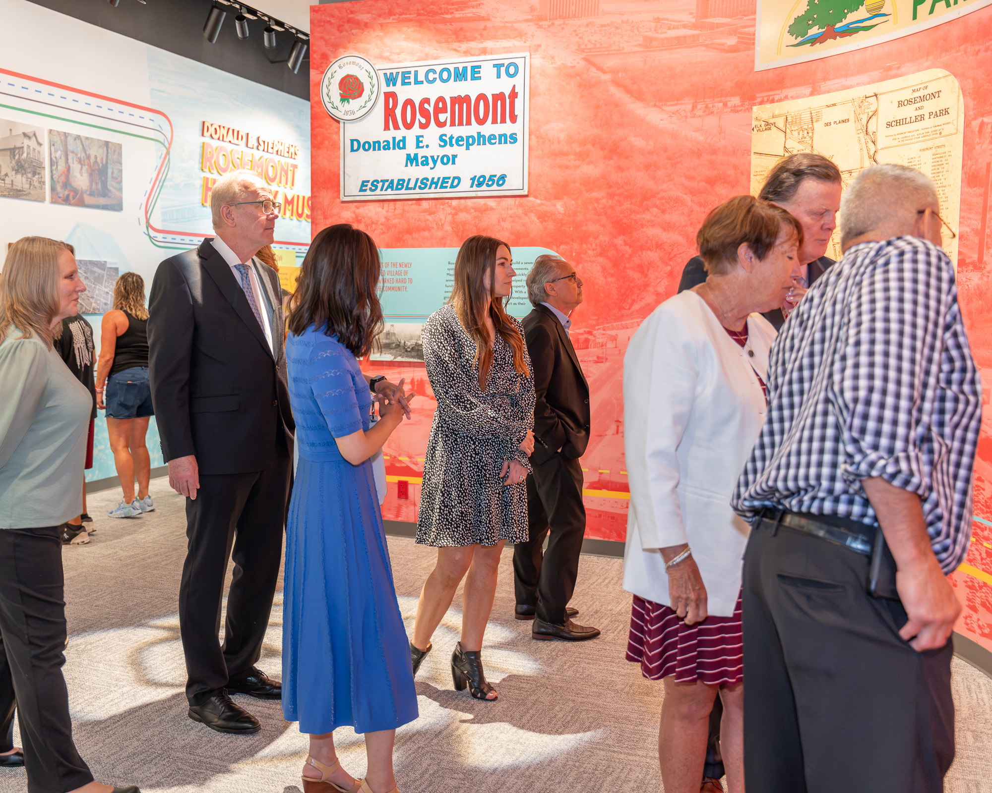 A group of people in semi-formal attire stand and observe displays in a museum with a large Welcome to Rosemont sign on the wall.