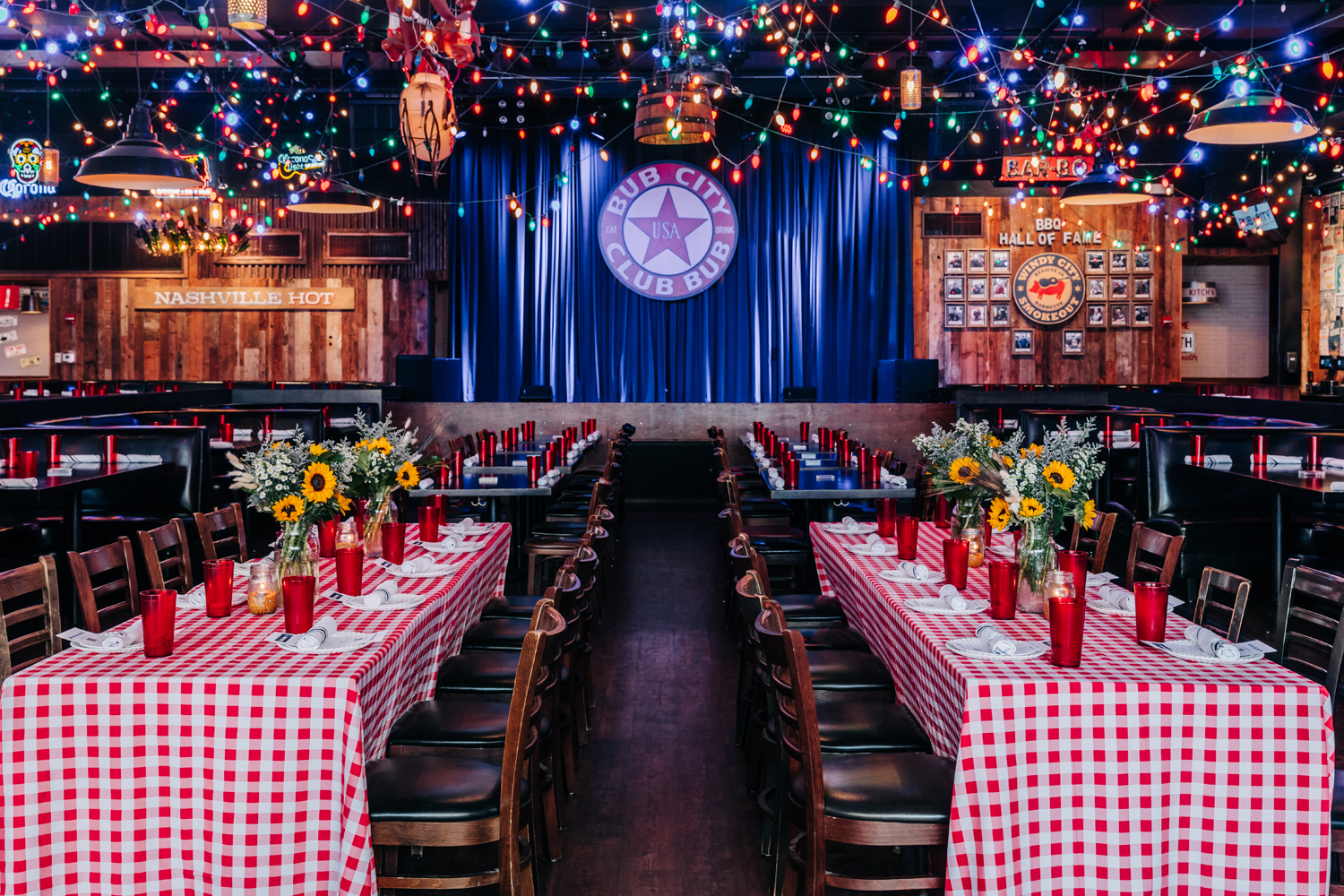 Dining room with rows of tables covered in red-and-white checkered tablecloths, each set with red cups, sunflowers, and black chairs, under a ceiling covered in multicolored string lights.