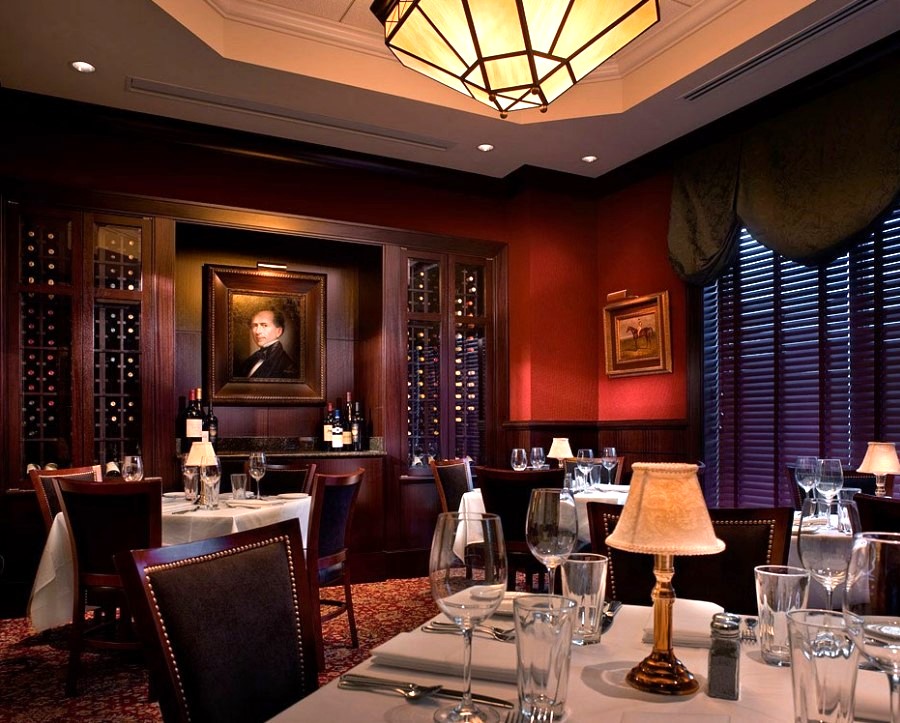 A dimly lit restaurant dining room with dark wood paneling, wine racks, set tables with white tablecloths, and a framed portrait on the wall.