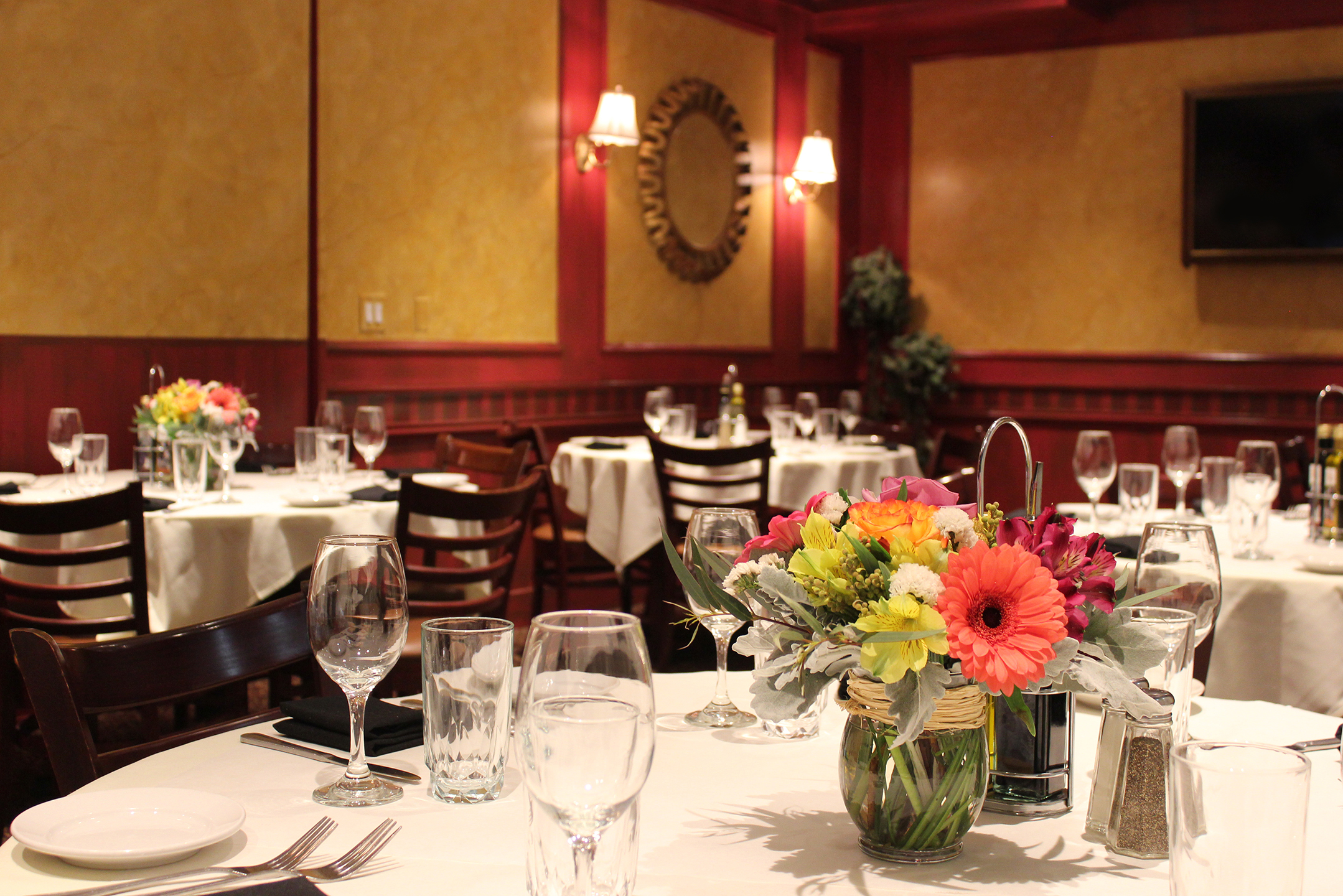 A restaurant dining room with multiple round tables set with white tablecloths, glassware, and flower centerpieces.
