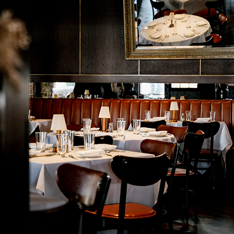 Dining tables with white tablecloths, glass tumblers, and small lamps arranged in a restaurant with leather booth seating and a wall mirror.