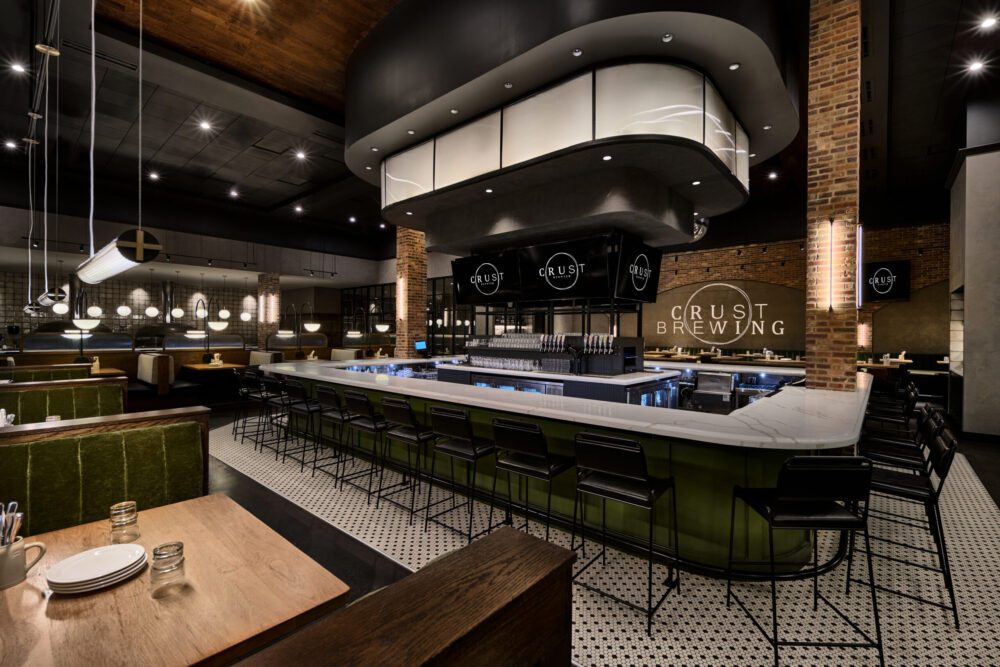 Interior of Crust Brewing featuring a large curved bar with stools, pendant lighting, and brick accents.