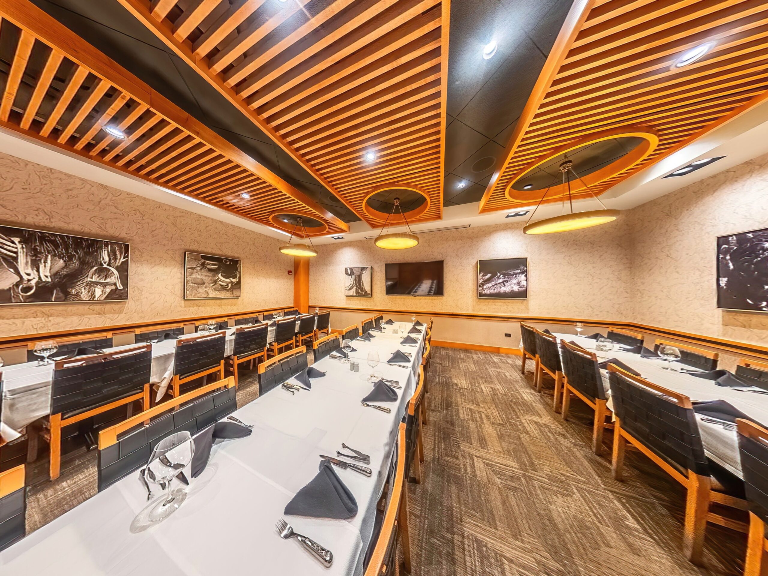 Restaurant dining room with long tables set for dinner, featuring wood-slat ceiling panels and black-and-white framed wall art.