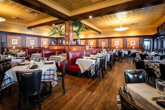 Restaurant interior with red leather booths, tables set with white tablecloths, and dark wooden chairs on hardwood floors.