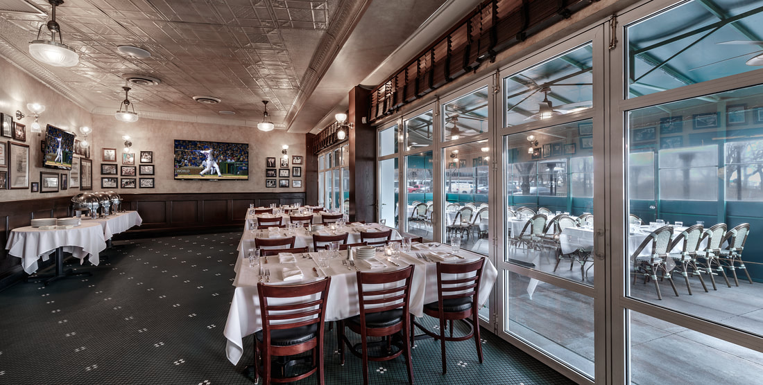An indoor restaurant dining room with white tablecloths, wooden chairs, and a TV on the wall, featuring glass doors that look out onto an enclosed patio with more tables.