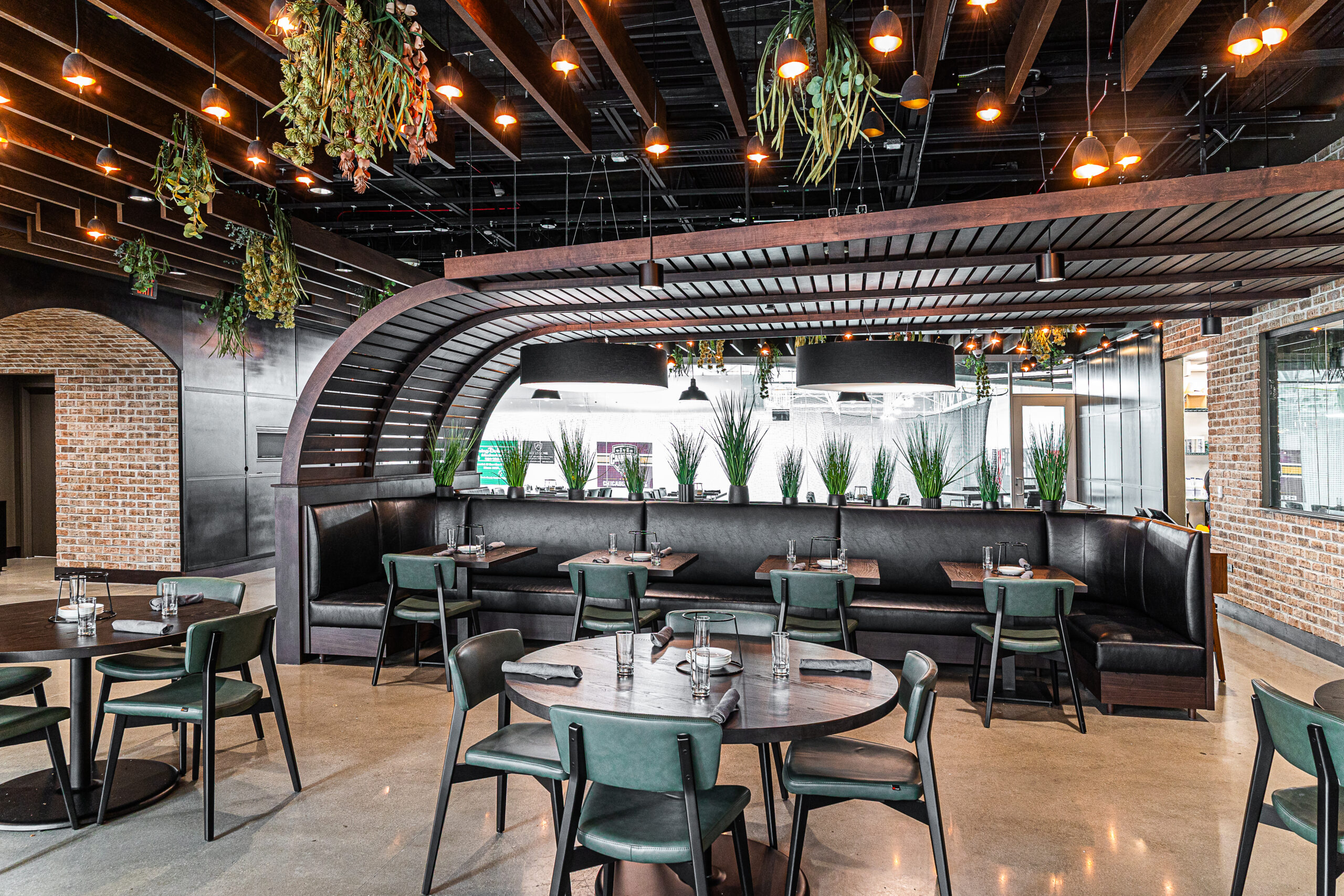 Restaurant interior with a curved wooden banquette, dark green chairs, round wooden tables, and hanging pendant lights.