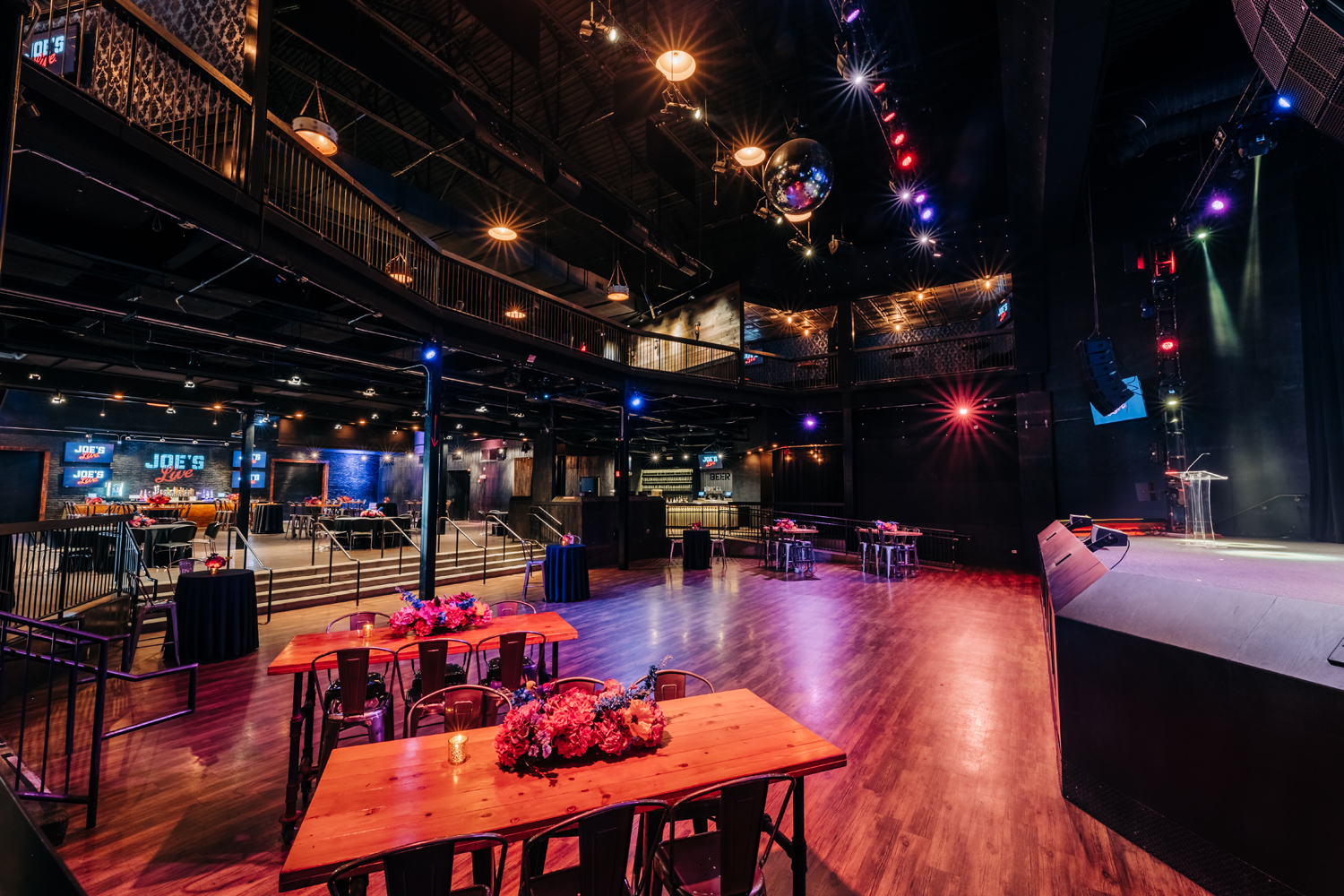 An empty event space with a wooden dance floor, a stage, and long tables set with floral centerpieces.