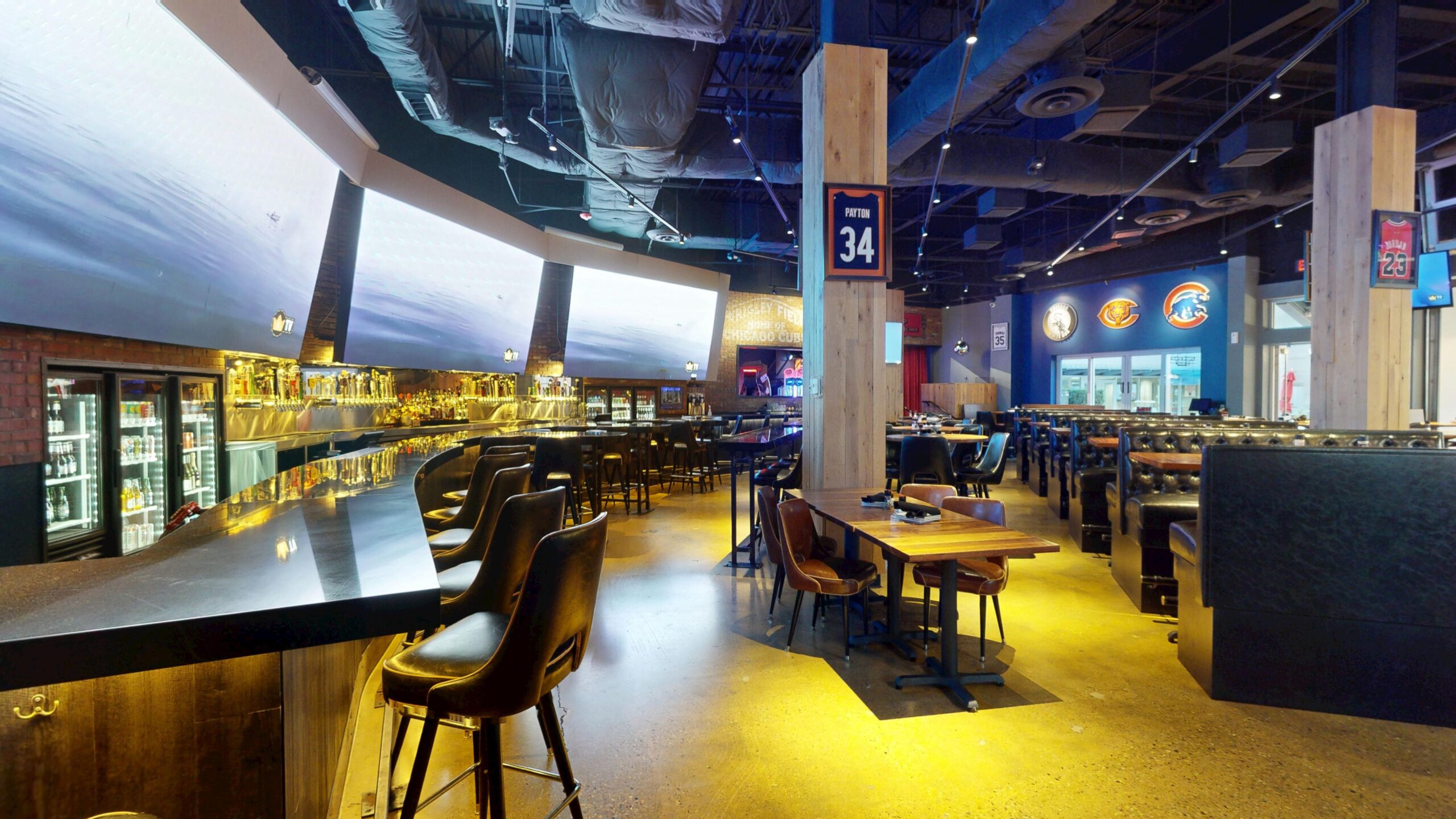 Interior of a sports bar with a long dark bar counter, high stools, dining tables, and Chicago sports team jerseys displayed on pillars.