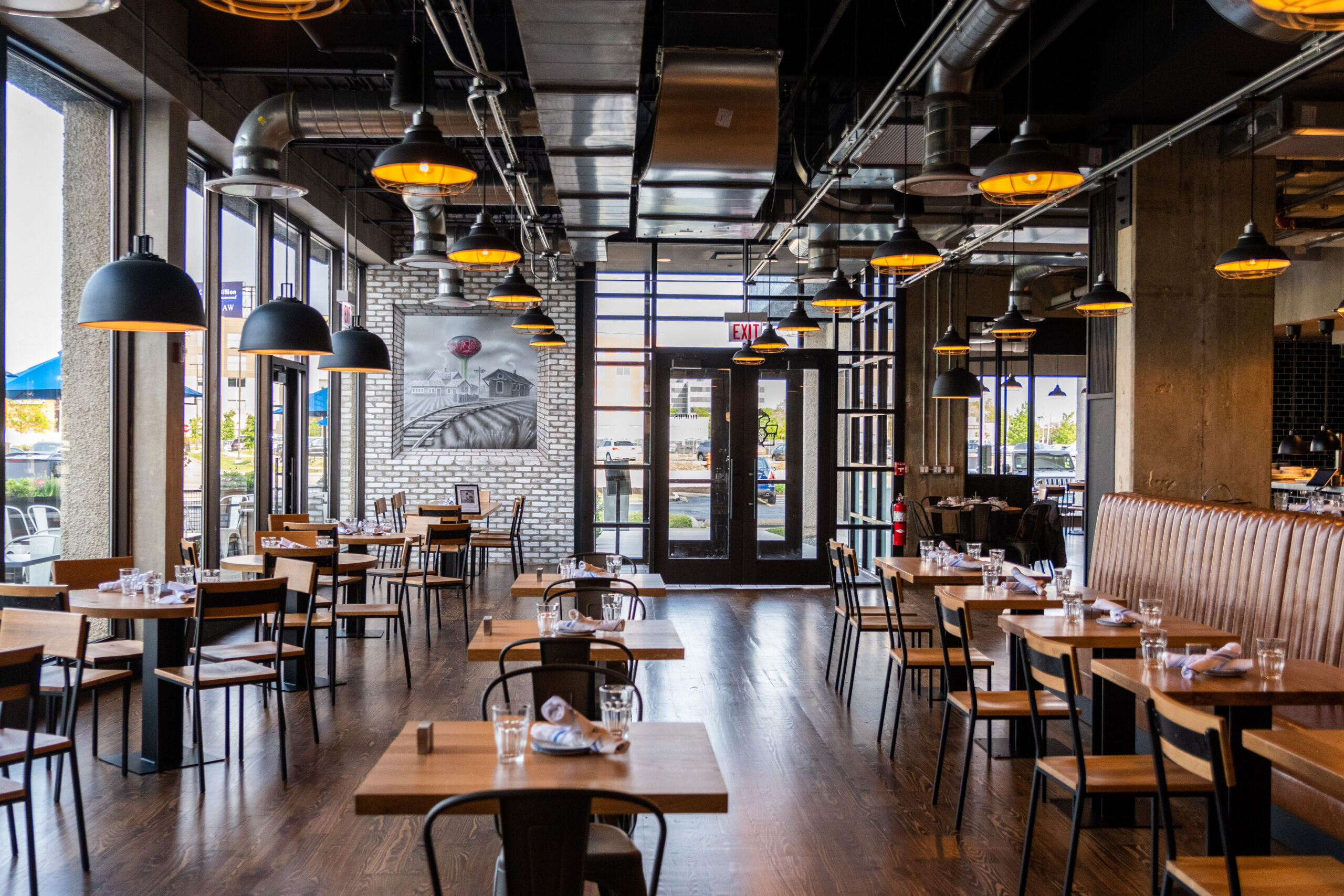 Interior of a restaurant with rows of empty wooden tables, black metal chairs, and hanging pendant lights.