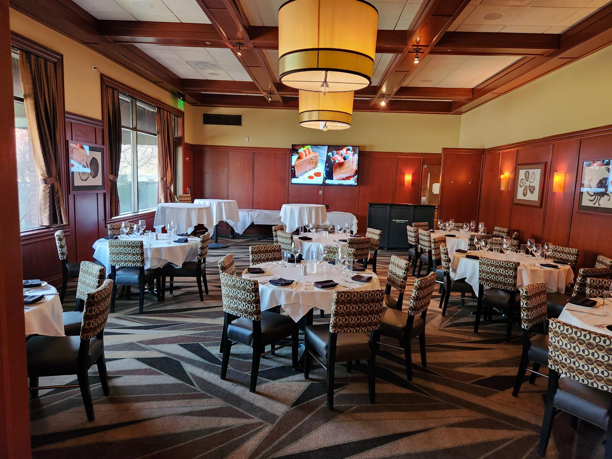 Dining room featuring round tables with white tablecloths, patterned chairs, wooden wall paneling, and two large hanging light fixtures.