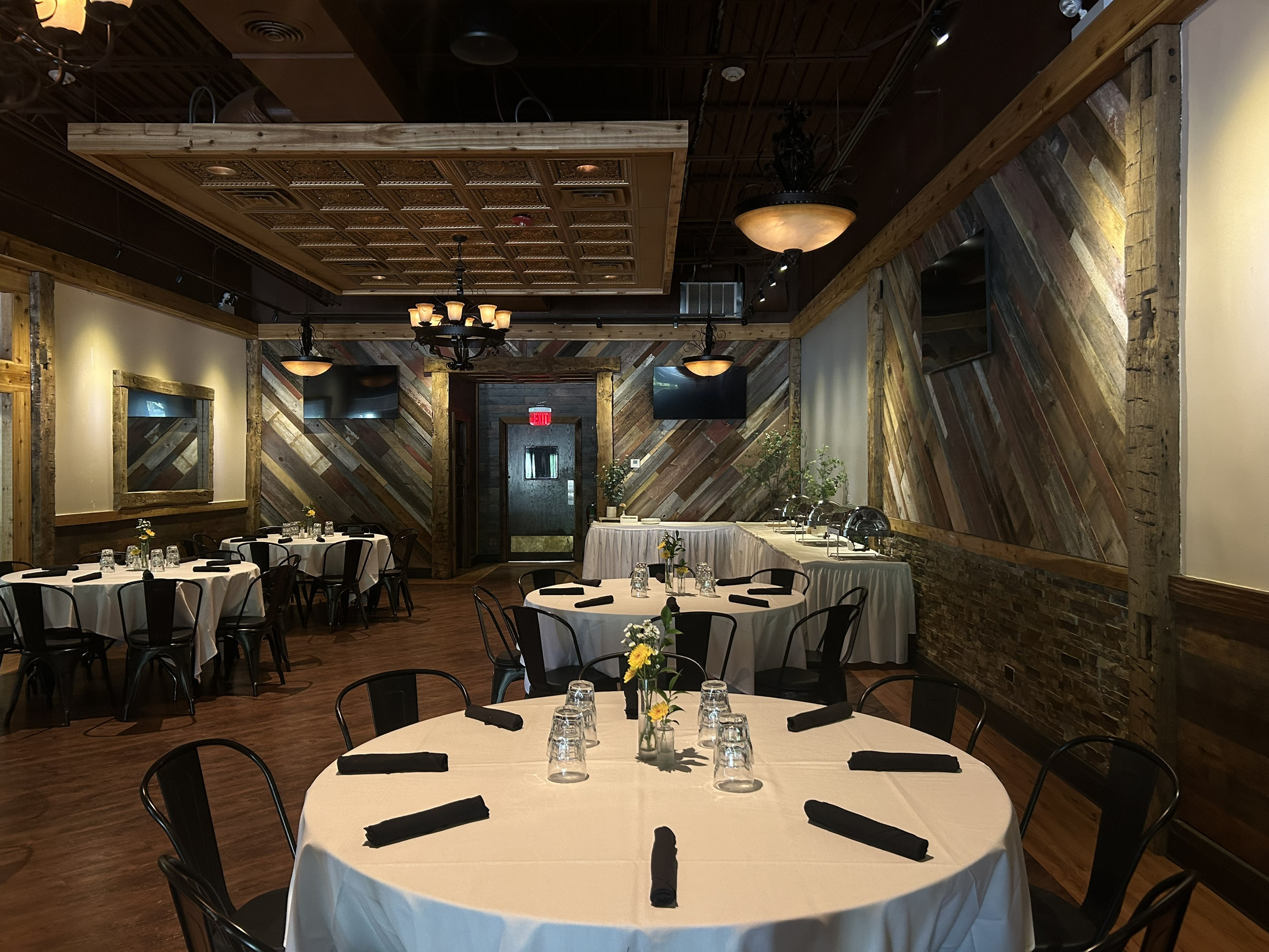 A restaurant interior featuring multiple round tables with white tablecloths, black chairs, and place settings, with rustic wood-paneled walls.