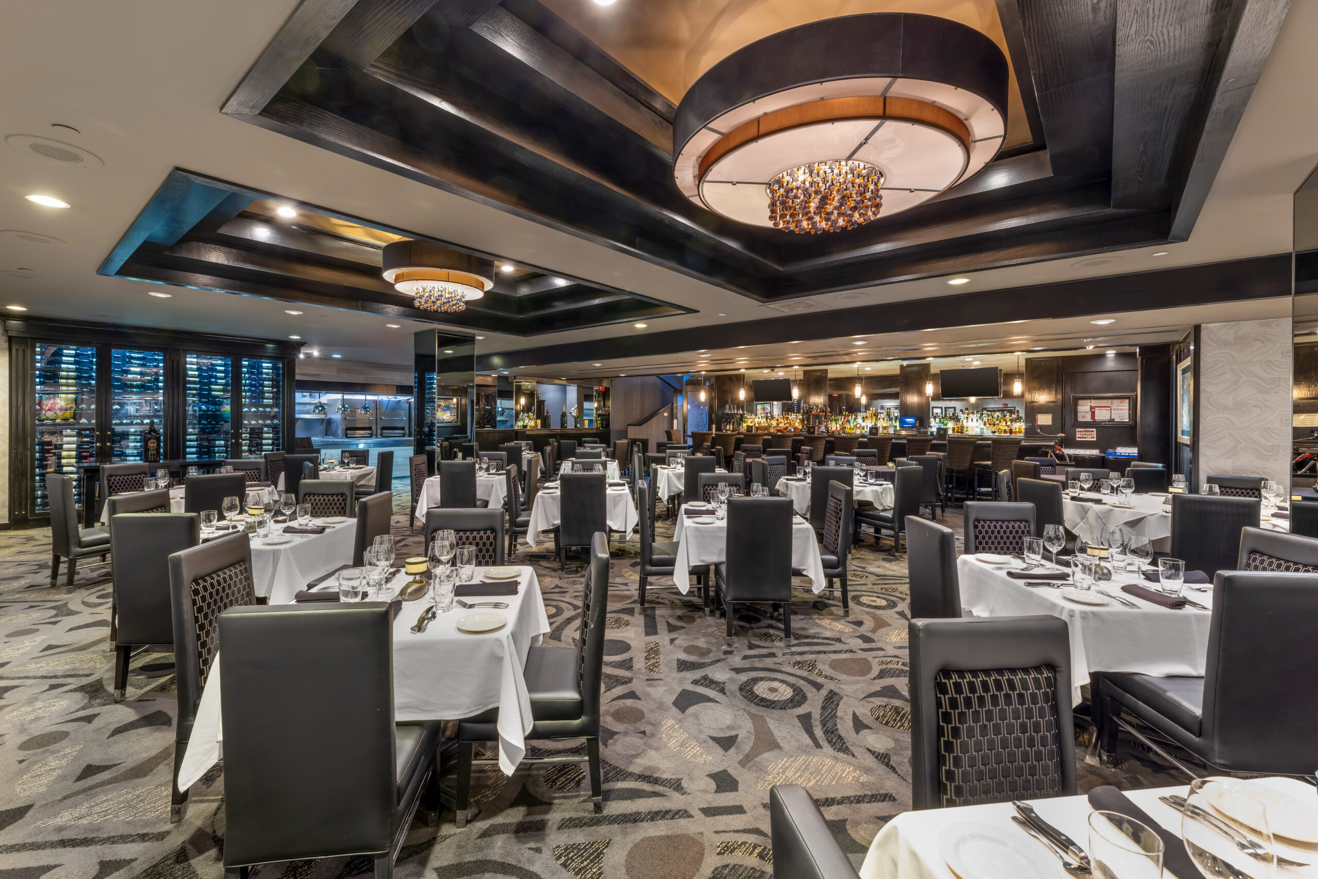 A restaurant dining room with white-clothed tables, dark upholstered chairs, and a bar in the background.