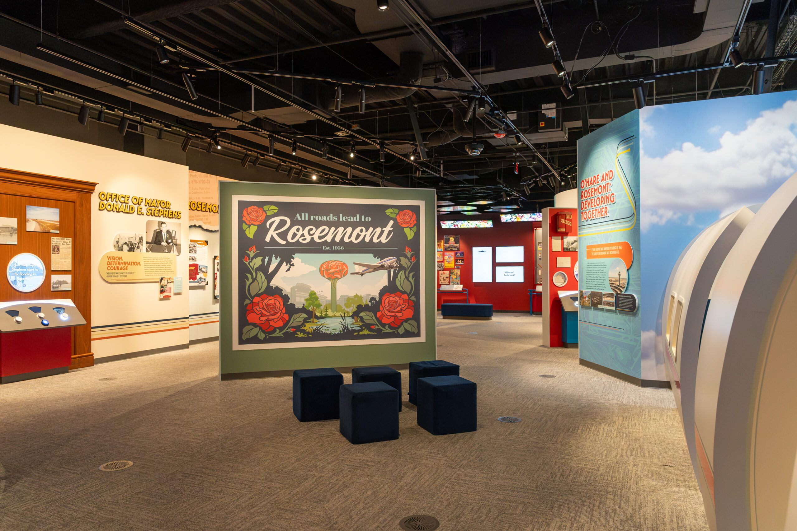 Museum exhibit interior featuring a central sign that reads All roads lead to Rosemont surrounded by red roses, with surrounding display walls detailing the history of Rosemont, Illinois.