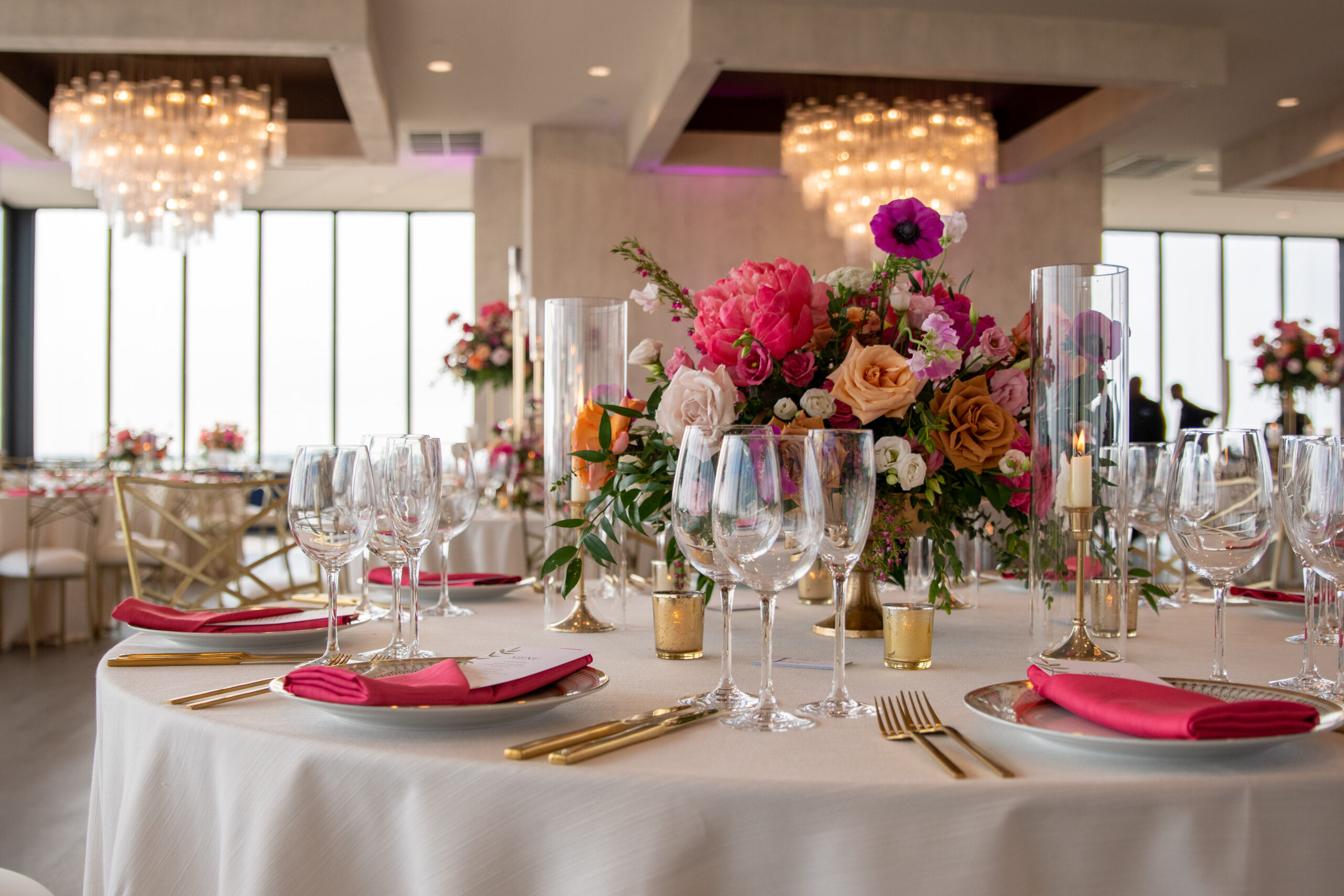A round table set for a formal event with pink napkins, crystal glassware, gold cutlery, and a large floral centerpiece.