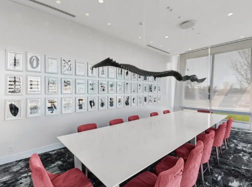 A conference room with a large white table, red velvet chairs, and a wall displaying a grid of framed abstract art prints.