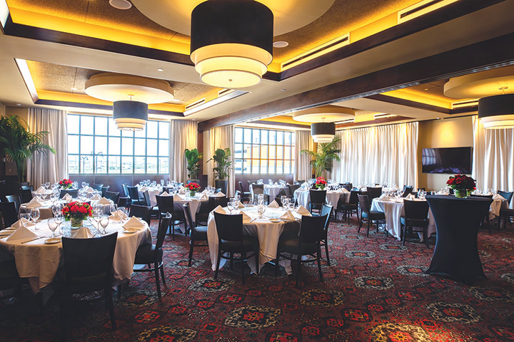 A banquet hall set with round dining tables, each featuring white linens, chairs, and centerpieces of red flowers.