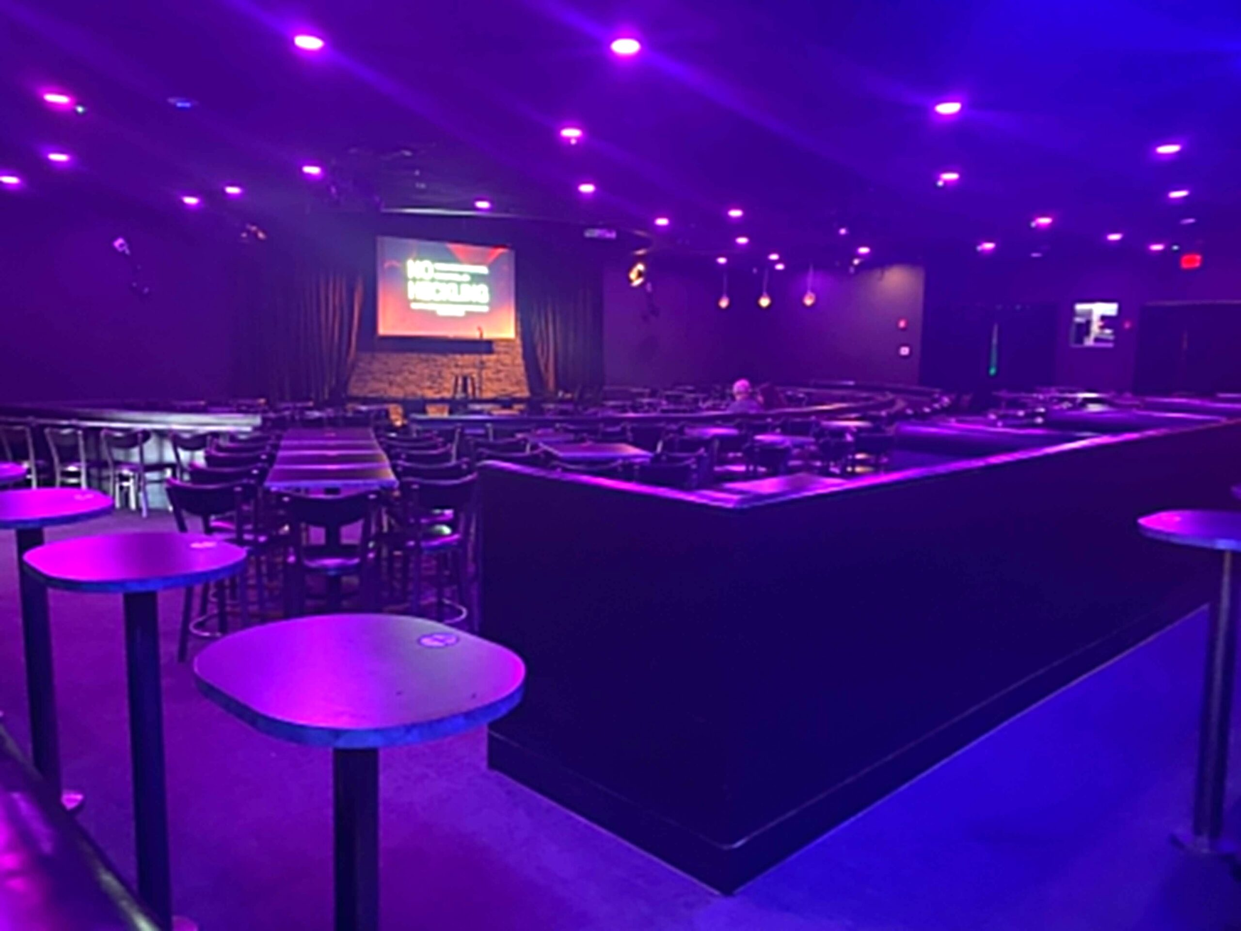 Interior of a comedy club with purple lighting, featuring a stage with a screen displaying No Heckling and rows of tables and chairs.