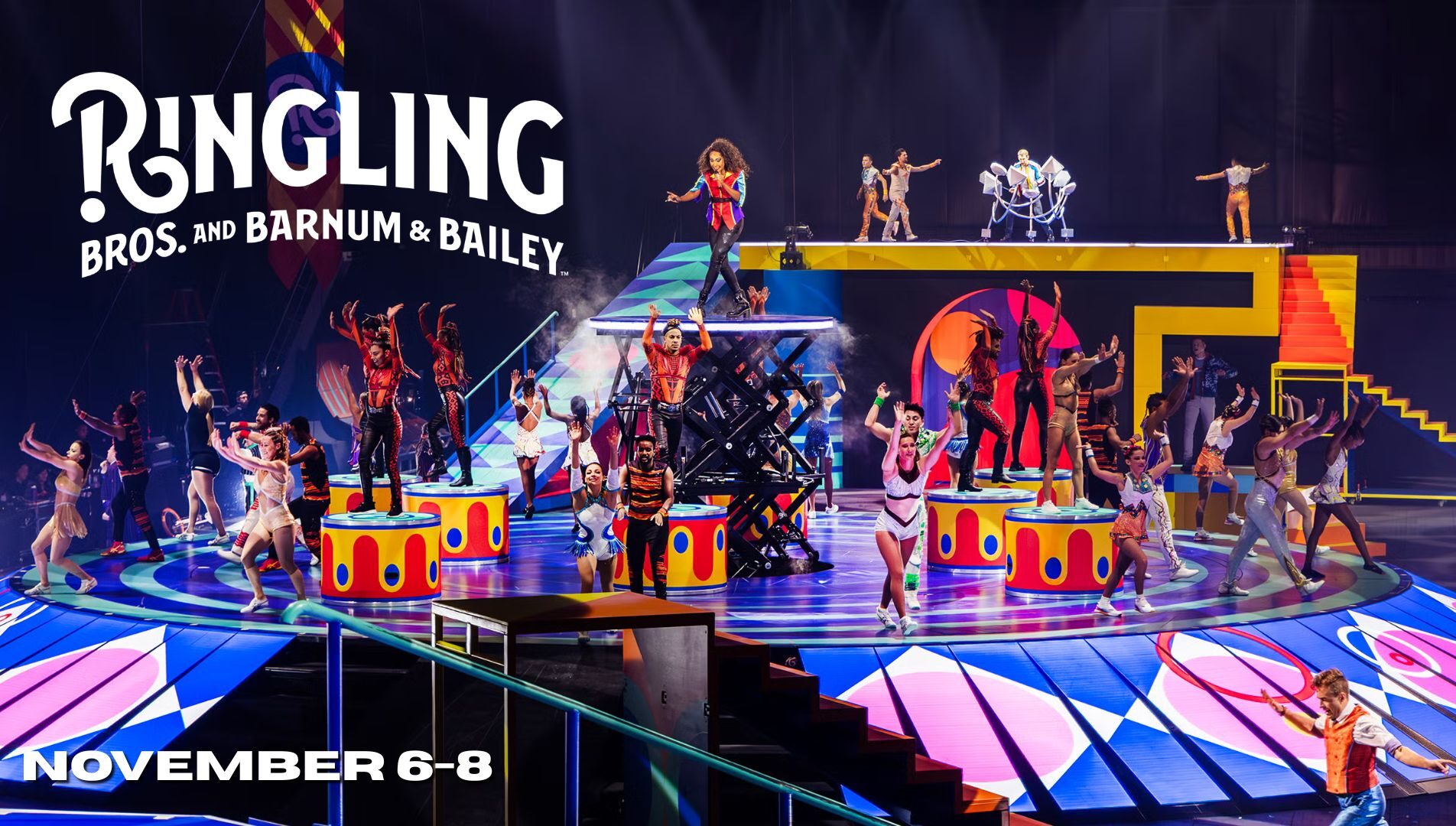 Circus performers in colorful costumes stand and pose on a multi-level, brightly lit stage with the text Ringling Bros. and Barnum & Bailey and November 6-8 displayed.